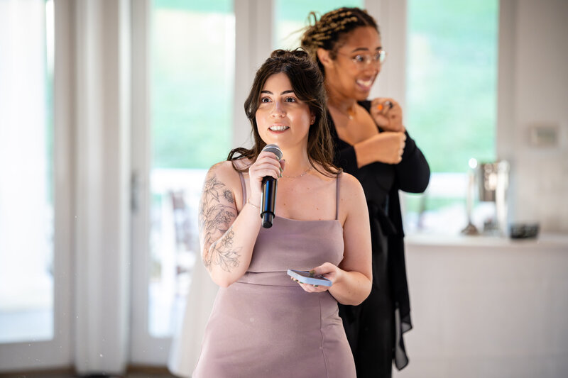 a petite white female in a spaghetti-strap mauve dress giving a speech with a microphone to wedding guests with a black female als interpreter signing out the speech dressed in all black 