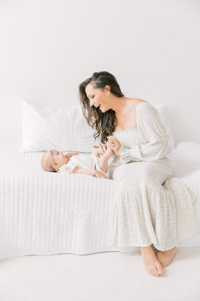 A happy mother in a floral print dress sits on a bed playing with her infant daughter's feet