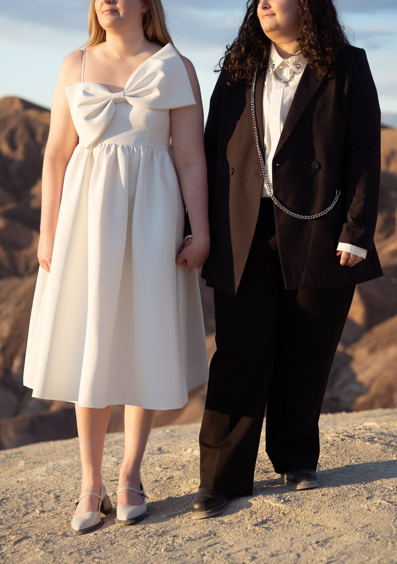 A lesbian couple holds hands and shows off their outfits. One woman is in a knee length white dress with a large bow, the other in a black suit with a silver chain detail