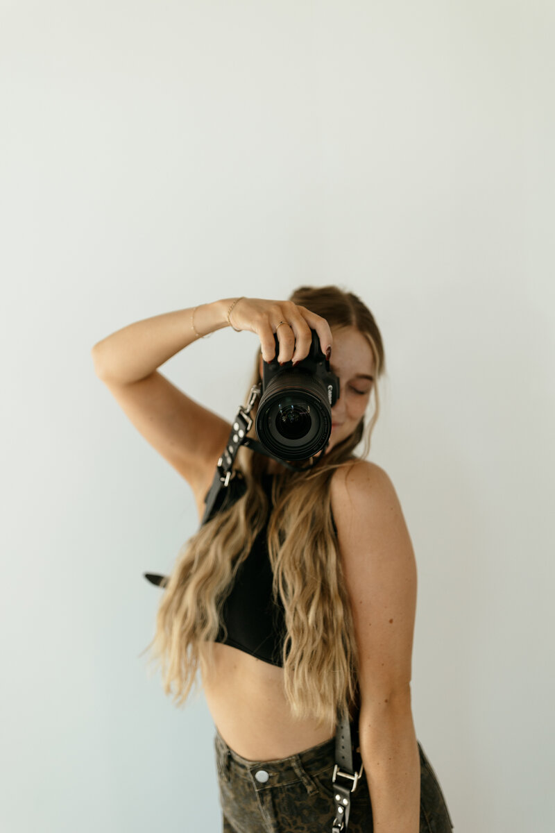 Blonde girl with a black hat posing on a stool with camera in her right hand