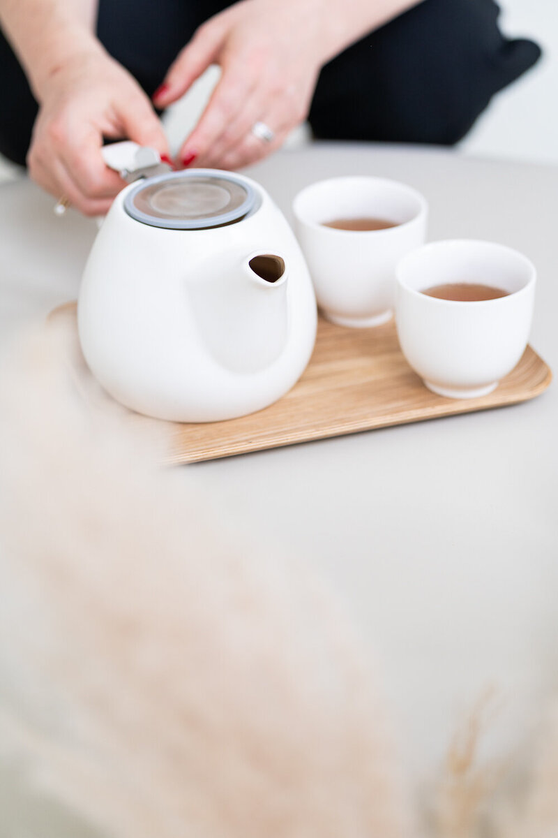 žena drží bílou keramickou konvici s čajem a vedle stojí šálky s čajem
