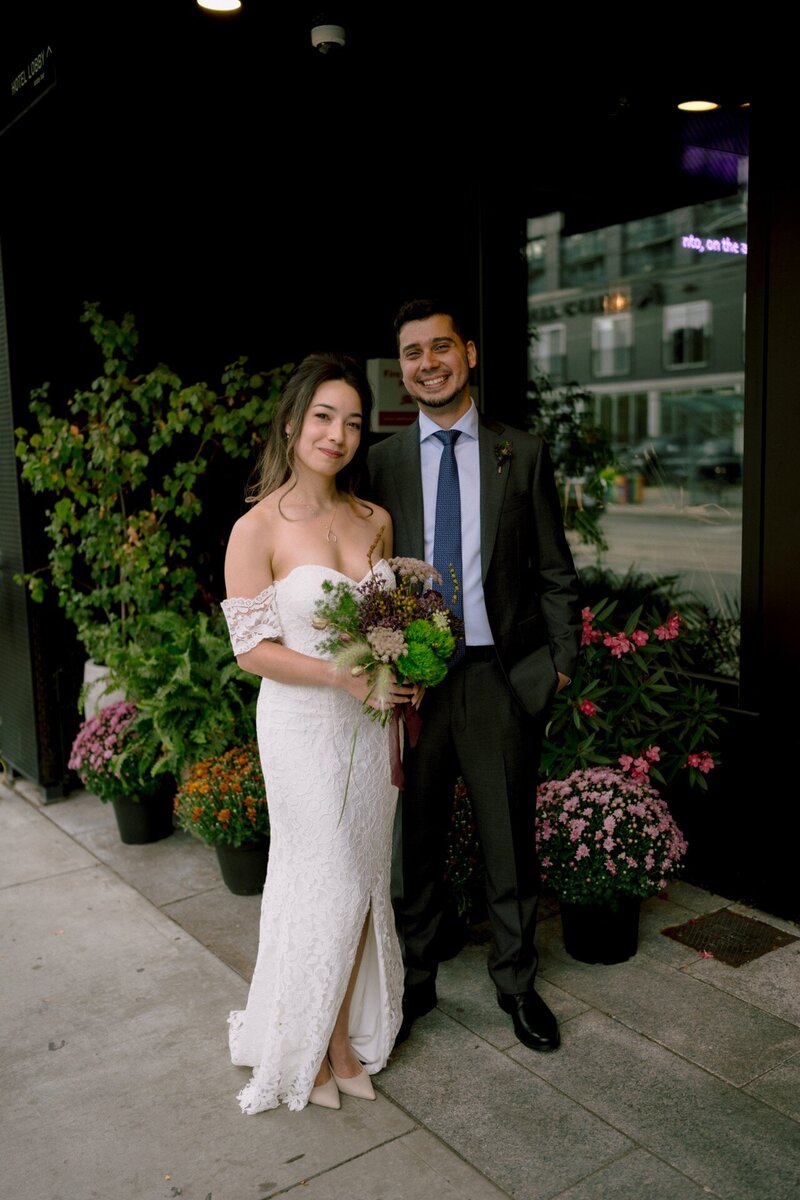 A couple dressed in wedding attire posing with a bouquet on a city sidewalk.