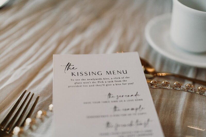 A minimalist white "Kissing Menu" with instructions sits on a gold-beaded charger plate on a table with a cream cheesecloth runner at a wedding at Le Belvedere in Wakefield Quebec, designed by Frid Events.