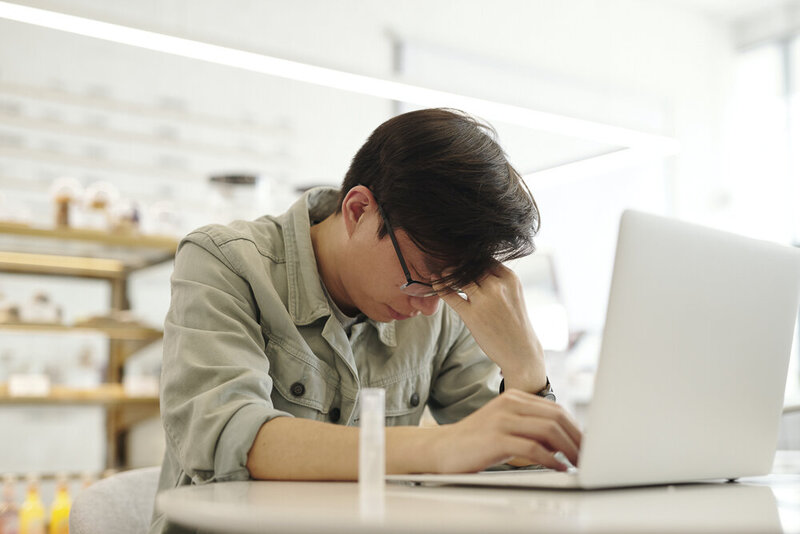 This image shows an Asian man wearing a light green button down and glasses, sitting at a desk, leaning over a laptop in front of him. His eyes are closed, and his face rests in his left hand with a tired expression on his face. Shelves are visible on the wall behind him.
