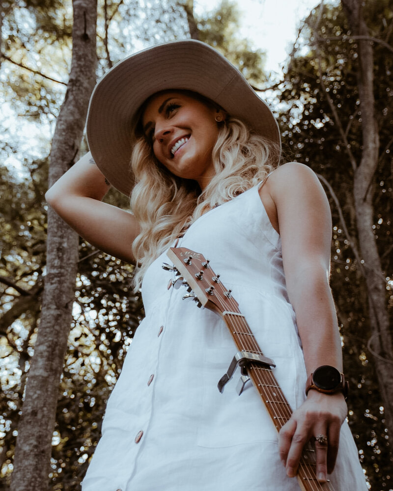 Blonde girl wearing a white dress  holding her brim hat and a guitar smiling in natural australian bushland