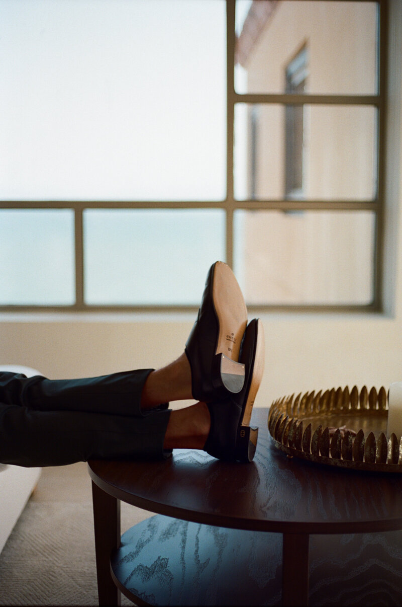 A person wearing loafers, has their feet up on a table, next to a window
