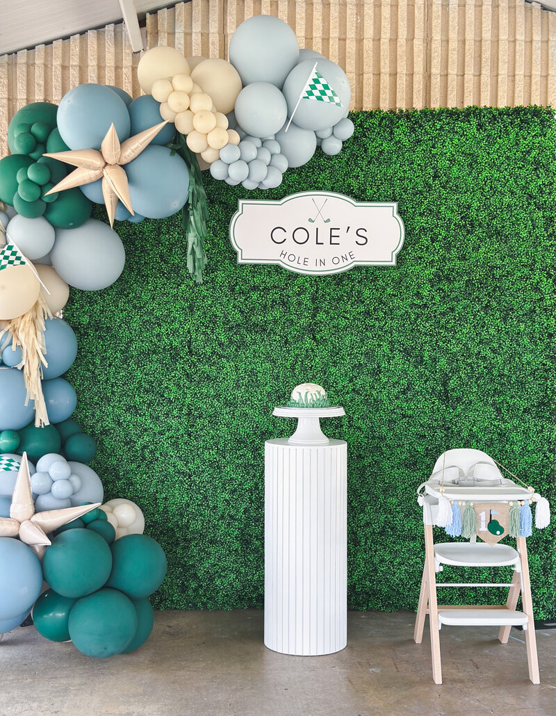 Golf balloon garland with green, blue and tan balloons hanging on boxwood floral wall with a custom sign and cake pedestal.