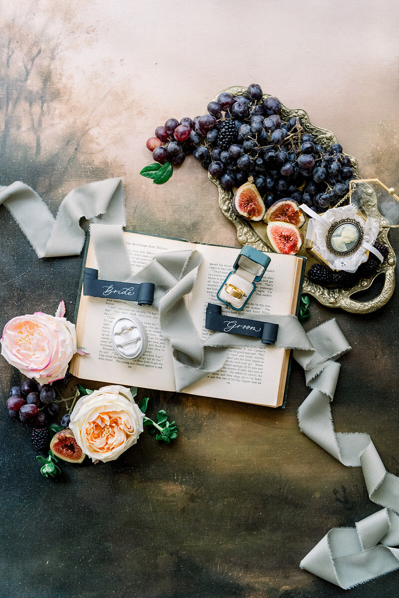 This stunning flat-lay arrangement captures the essence of timeless elegance with a vintage-inspired styling. A combination of ripe grapes, figs, and vibrant blooms surrounds an open vintage book with calligraphy cards labeled “Bride” and “Groom.” The textured velvet ribbons and antique tray add a luxurious touch, while the engagement ring sparkles amidst the rich setting. This California wedding-inspired photo radiates sophistication and charm, perfect for couples seeking a refined, romantic theme for their big day.