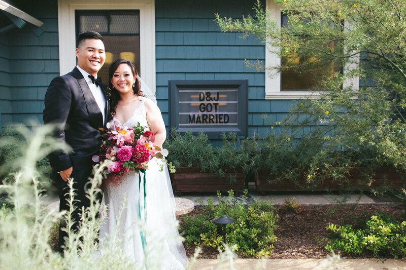Bride and groom embrace and smile brightly outside blue shingled house on wedding day