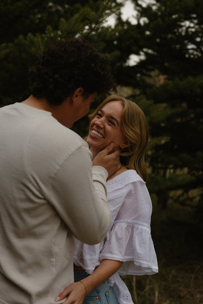 Couples photography in Rexburg, Idaho Falls, and surrounding areas. A man is holding his girlfriend over his shoulder, walking through a grassy field with trees in the background.