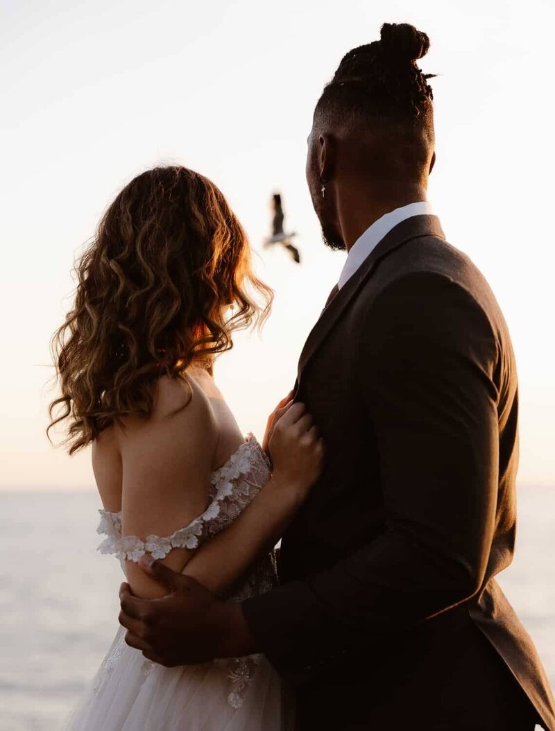 A couple stands close together at sunset, looking out toward the ocean in Big Sur. The bride wears an off-the-shoulder dress and the groom is in a suit, with a bird flying in the background.