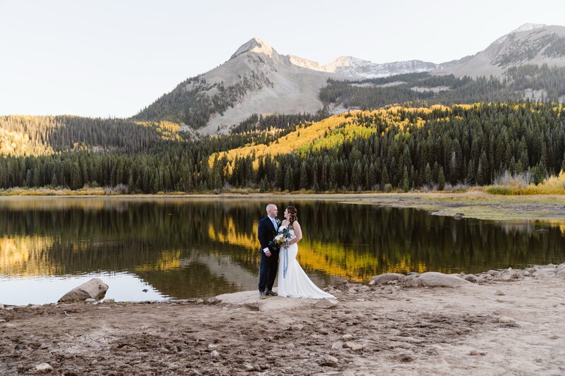 Couple standing by a reflective alpine lake surrounded by golden fall aspens during their Crested Butte Colorado elopement, captured by a Colorado elopement photographer and videographer Alpine Vows.