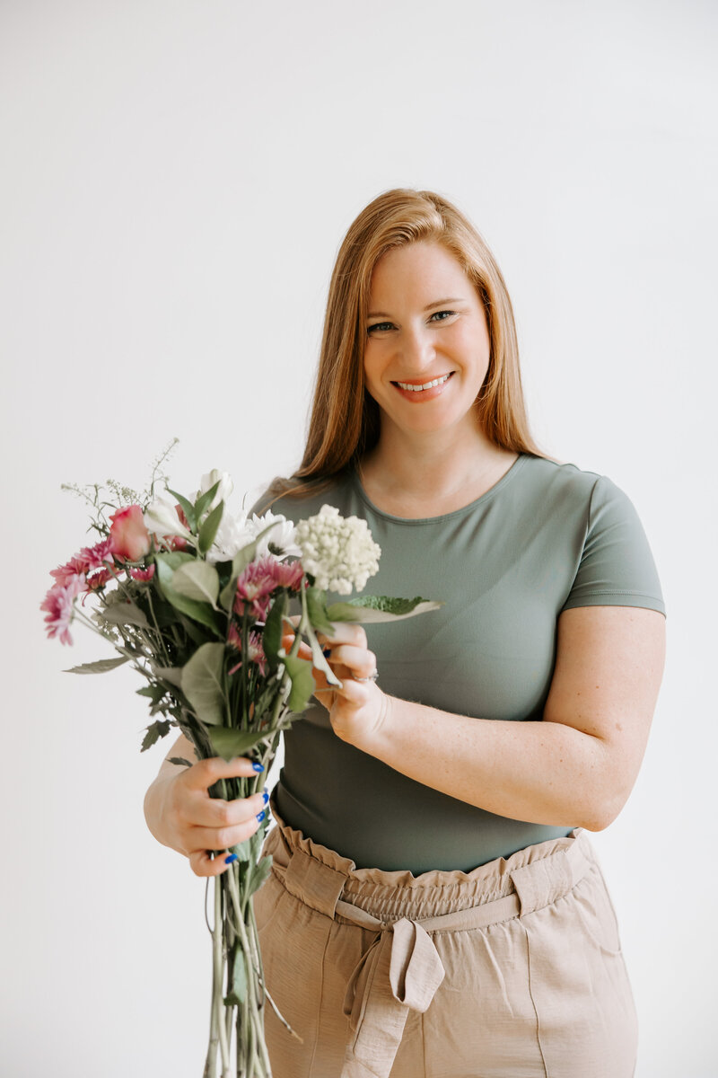 Alicia stands with a bouquet of fresh pink and white flowers, wearing a sage short sleeve shirt and loose khaki pants that tie at the waist.