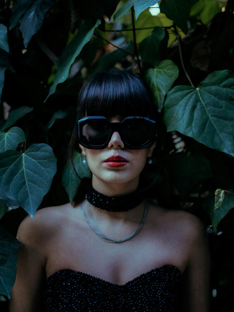Dark image of a fierce woman with sunglasses and a black dress against a tree.
