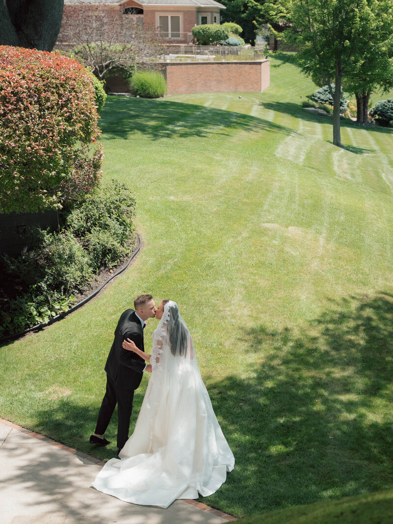 Bride and groom kissing on sunlit lawn with full ballgown and dramatic veil
