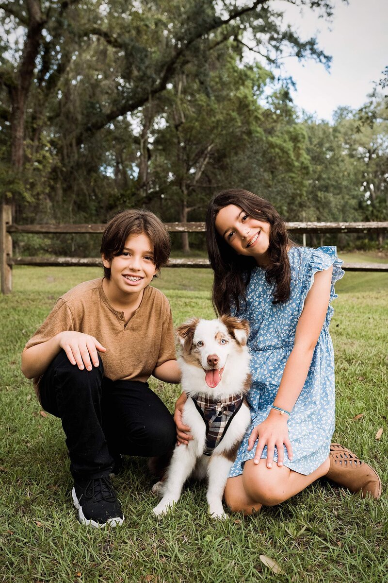 A boy and girl smile while posing with their dog in a sunny park setting.