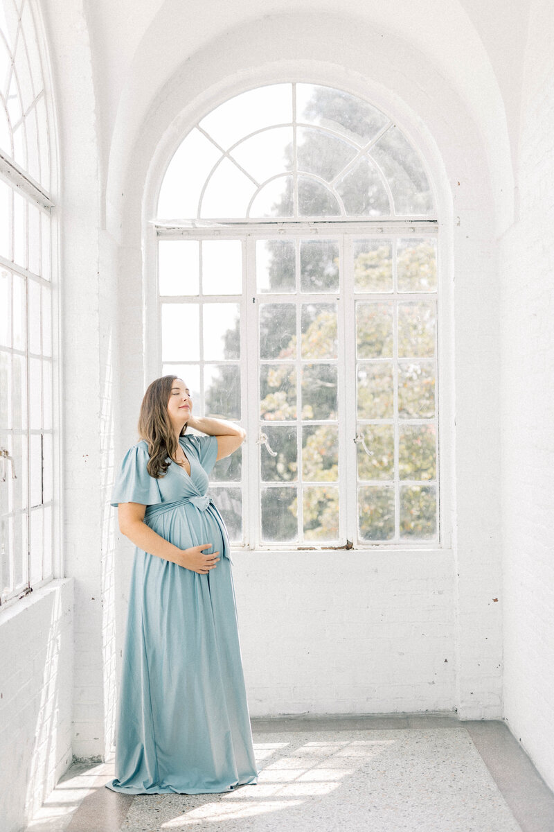 luxury knoxville mom wearing blue dress stading in huge white hall in the sunlight by the window at the bleak house