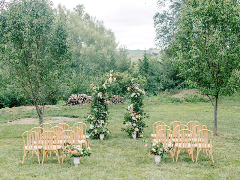 Wedding ceremony decor with green, pink, and white floral arch, potted vases, and wooden chairs overlooking a green vista