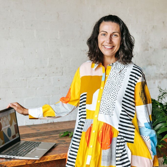 A woman with dark, shoulder-length hair stands next to a table with an open laptop. She is wearing a vibrant, patterned dress with a mix of colors including yellow, orange, and black. The background is a neutral, textured wall.