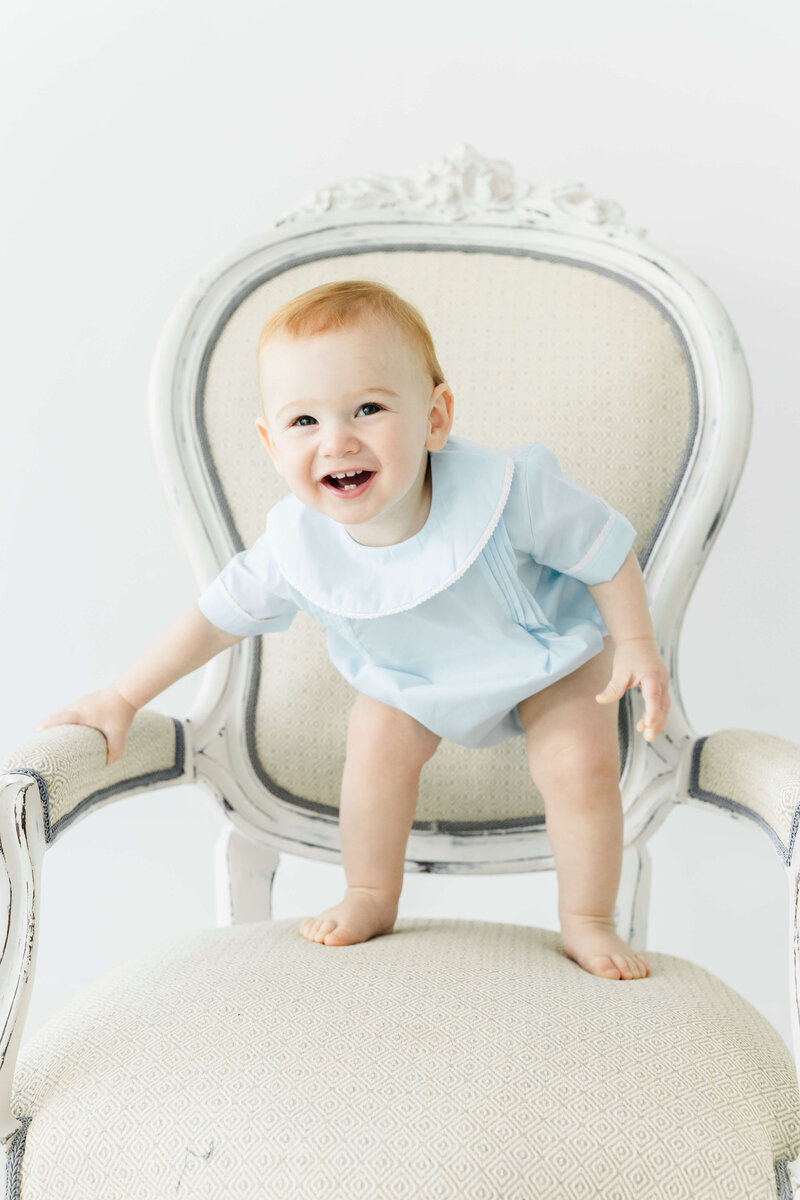 Baby standing on a chair smiling at the camera wearing heirloom outfit