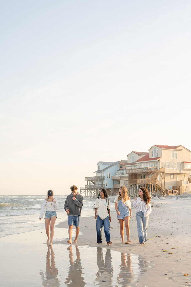 five friends walk barefoot on the beach with beach houses in the background during golden hour