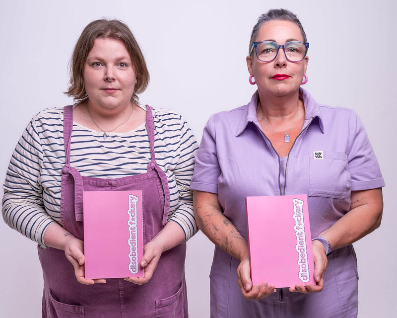 Lucy, in a striped top and purple dungarees, and Pippa, in a lilac jumpsuit, stand side by side holding pink “Disobedient Fuckery” notebooks in front of them during a Disobedient Business® Co. photoshoot. Both face the camera with calm, serious expressions, kinda 'The Shining' vibes.