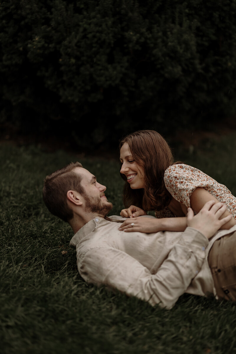 A couple lies on a grassy field, smiling at each other. The woman, in a floral dress, leans over the man, who wears a light-colored shirt. Captured by a New England Wedding Photographer, they share a joyful moment amidst the lush greenery.