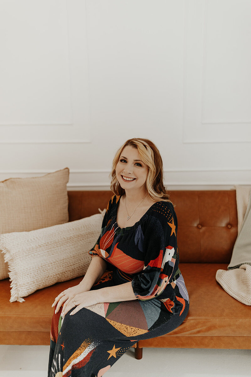 Shylah sits on a brown leather couch, smiling toward the camera, hands in her laptop. She is surrounded by cozy pillows, and she is wearing a brightly-colored patterned dress.
