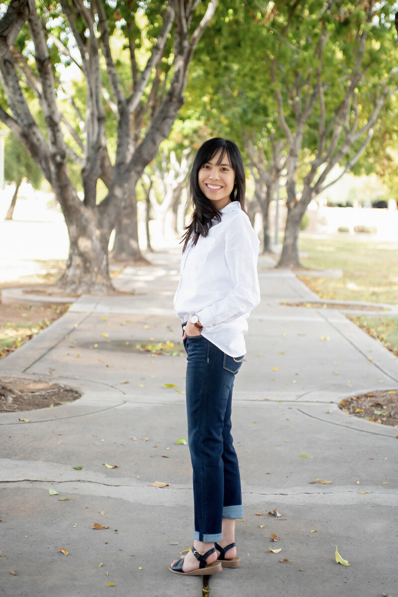 Laci walks down a park path and turns back and smiles, wearing a white blouse and jeans