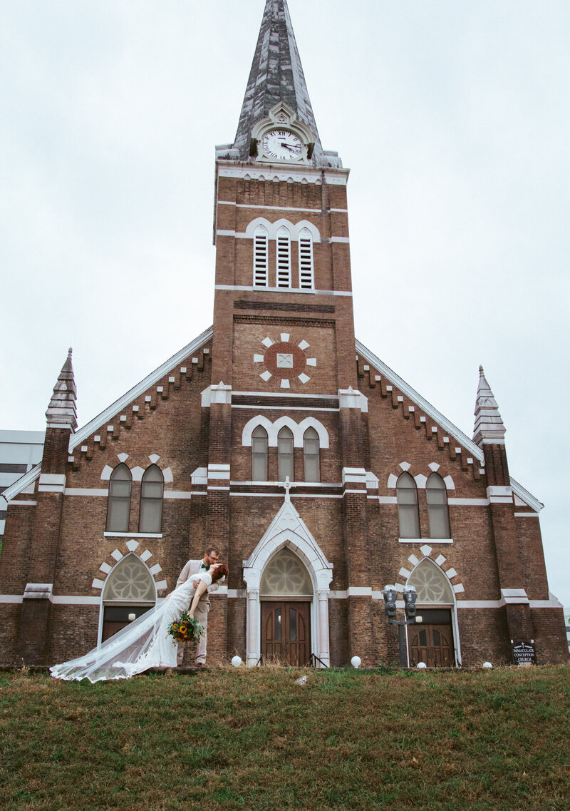 groom kissing bride on grass in front of large church