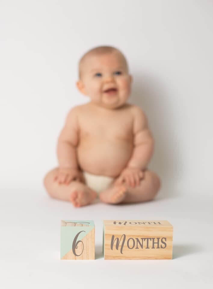 A smiling six-month-old baby sits in a diaper with wooden blocks reading "6 Months" in front of them.