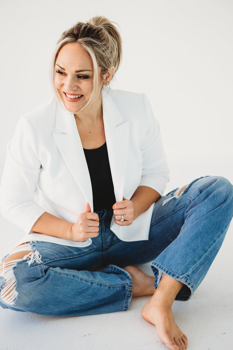 Woman wearing a white blazer and jeans is sitting on the floor and smiling in another direction