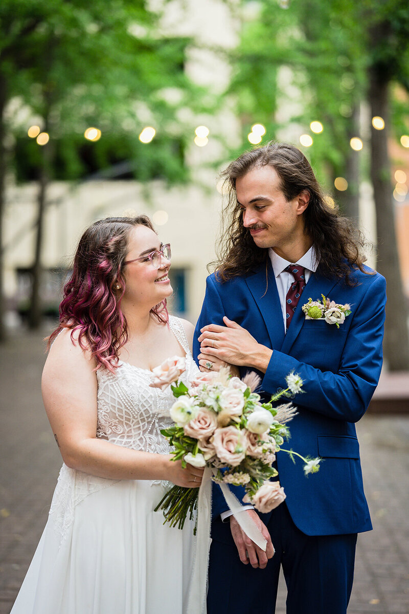 A bride and groom stand together for a photo during their elopement day in Downtown Roanoke in Virginia. The pair look at one another and smile as the bride holds out her bouquet slightly in front of them.