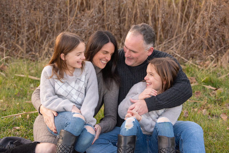 A family of 4 sitting down and holding onto each other and smiling at their family photoshoot.