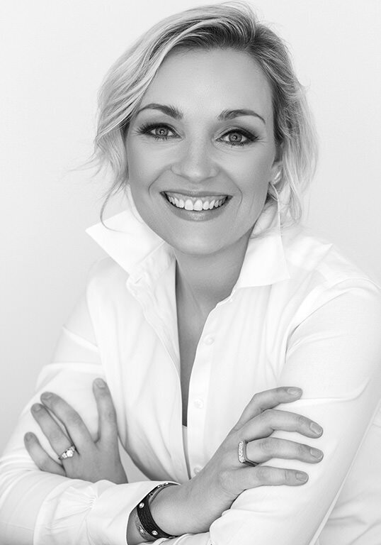 A professional studio portrait of a smiling blonde woman in a crisp white button-down shirt, with elegant jewelry, looking confident.