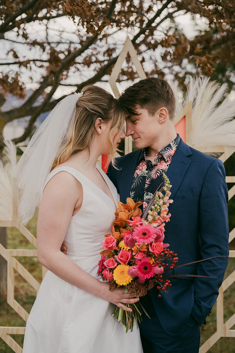 Close up of bride and groom outside, in front of a bespoke ceremony backdrop. They are close together with their foreheads touching. The bride holds a colourful bouquet and the groom wears a colourful shirt with his blue suit.