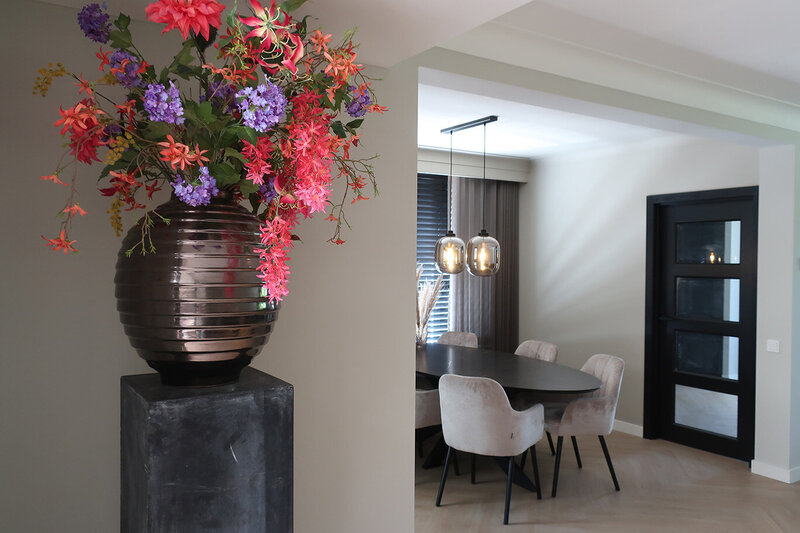 Grote vaas met kunstbloemen in felle kleuren op sokkel in strak interieur met eethoek op de achtergrond