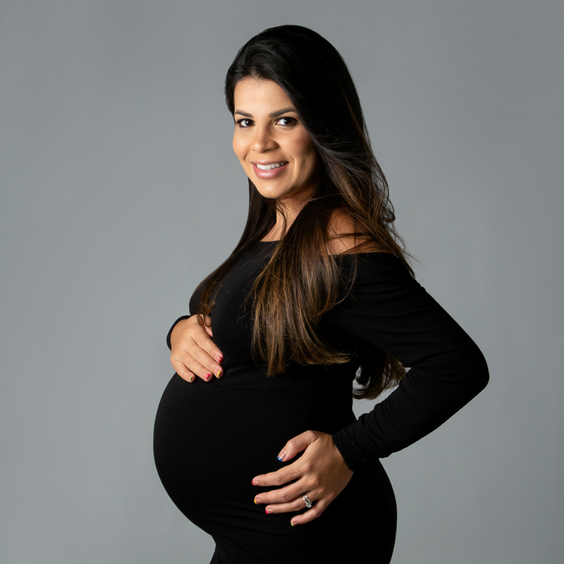 simple pregnancy photo of charlotte nc mom to be Gabbie wearing a black dress on a grey background long brown hair smiling at the camera with her hands on her belly emphasizing her pregnancy.