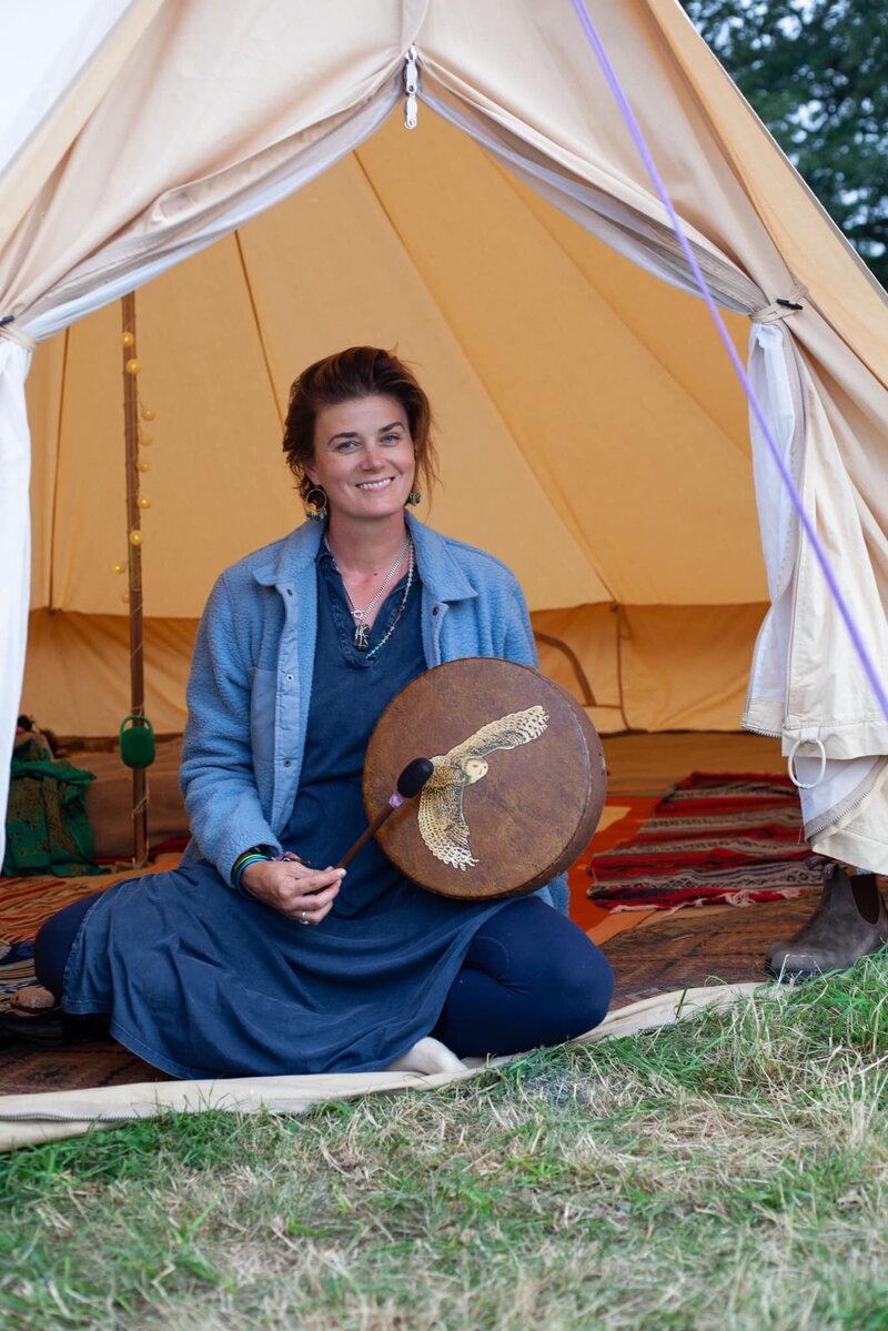 A sound healing practitioner from Otrain Wellness sits outside a ceremonial tent in Tulum, holding a sacred drum adorned with a golden eagle, ready to begin an intuitive energy healing session.