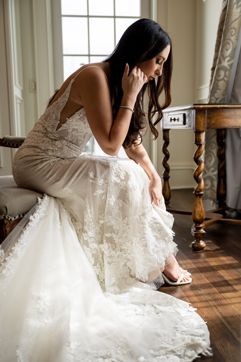 Bride sitting in front of a window holds her hair back as she reaches down to fix her shoe.