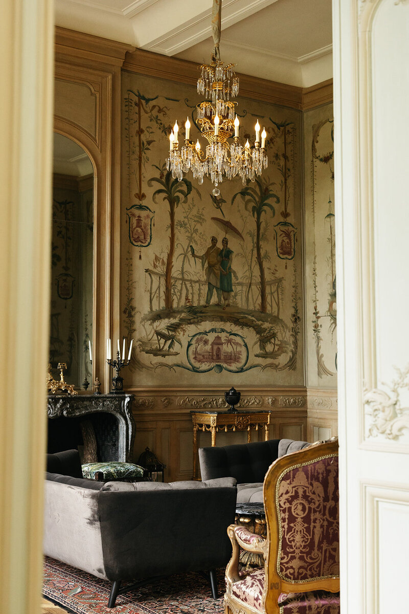 A luxurious French chateau interior featuring a fireplace, painted walls with intricate murals, and elegant chandeliers