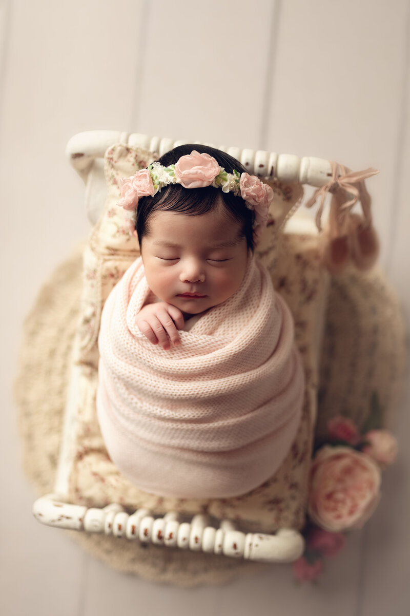 Newborn girl styled with pink wrap and floral crown, posed on a delicate vintage bed setup.
