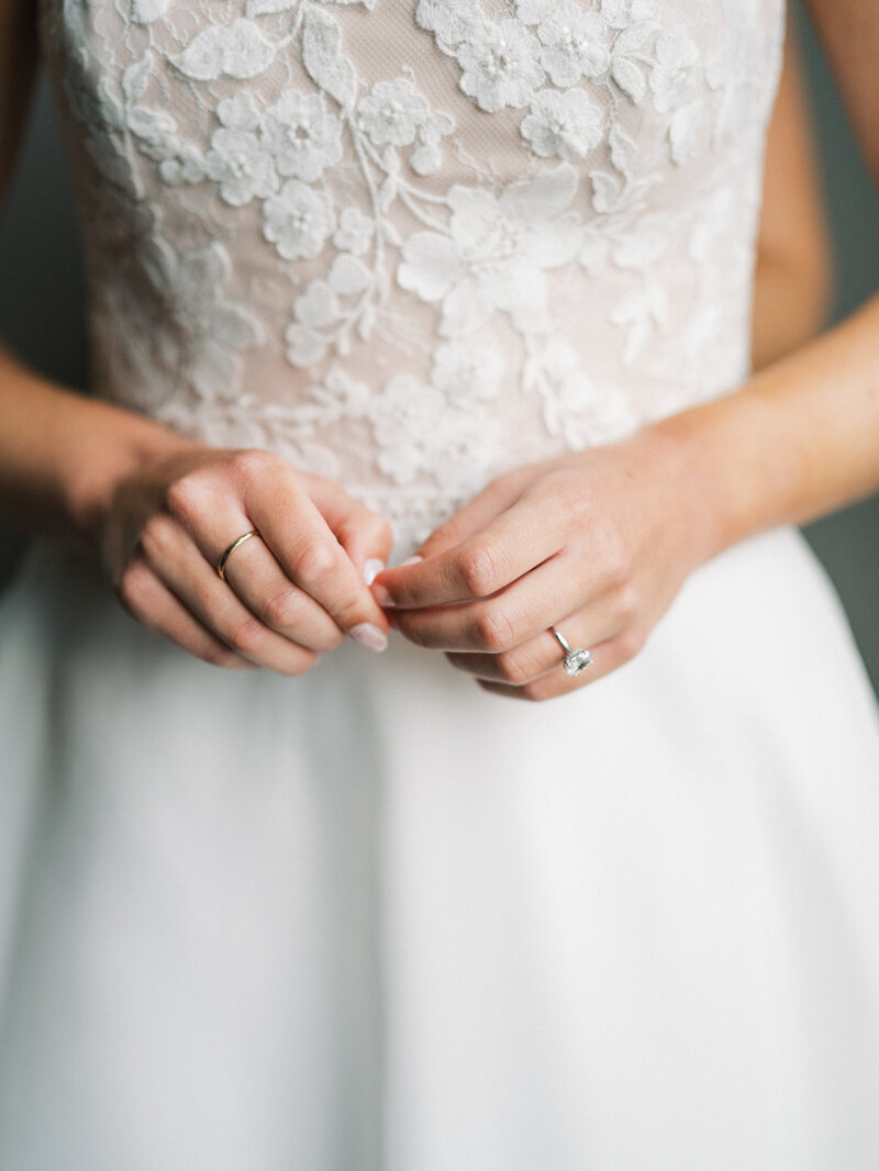 Close-up of a bride’s hands holding her dress, showcasing delicate lace details and her engagement ring."