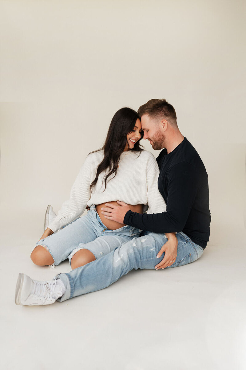 Husband is hugging his pregnant wife while there are both sitting on the flor  in the photo studio