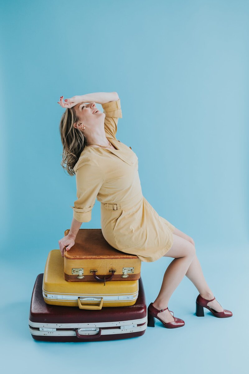oman with shoulder-length blonde sits atop several vintage suitcases with her hand over her forehead in front of an aqua backdrop.