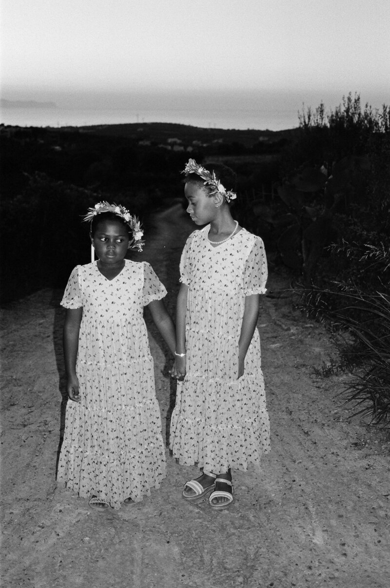 Two African American sisters dressed as flower girls at a wedding in Greece, sharing a joyful moment in elegant dresses.