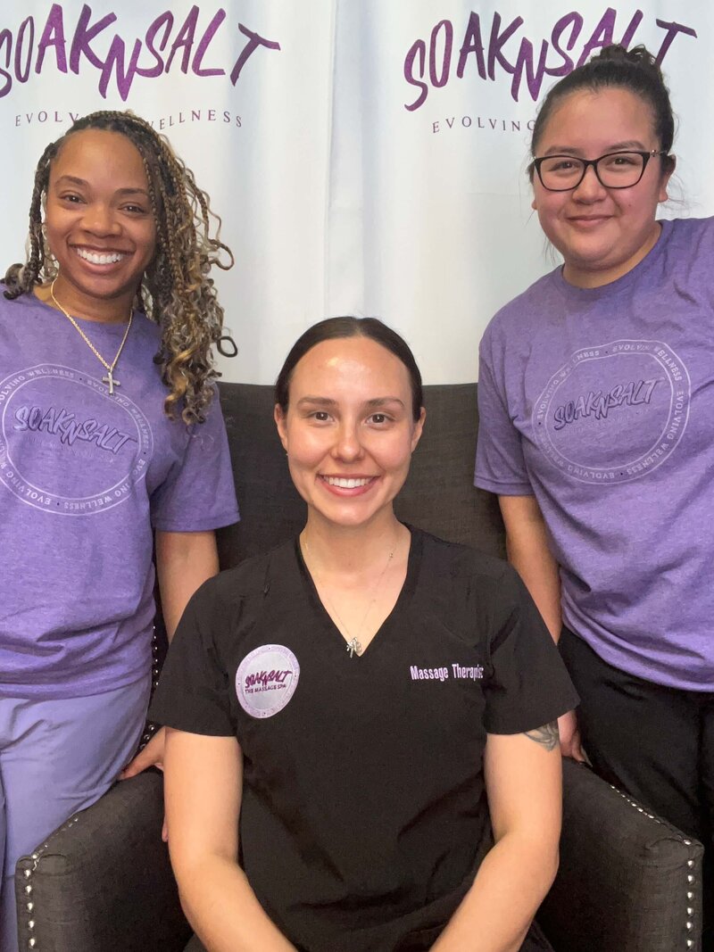 Three women smile at the camera in front of a “SoakNSalt Evolving Wellness” backdrop. Based in Tolleson, Arizona, they proudly represent massage therapy, wearing logo shirts and a black massage therapist uniform.