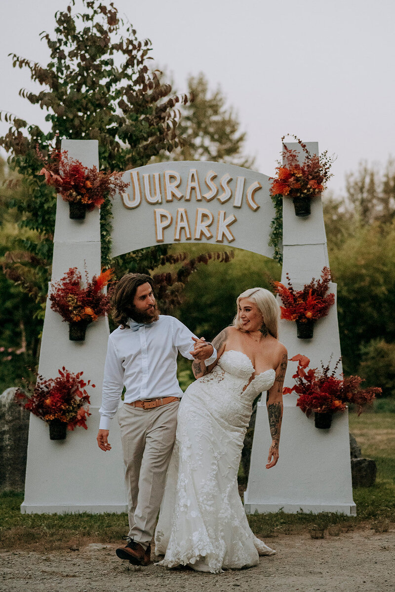 A wedding couple bump hips in front of a Jurassic Park sign at their wedding at Cheekeye Ranch, Squamish