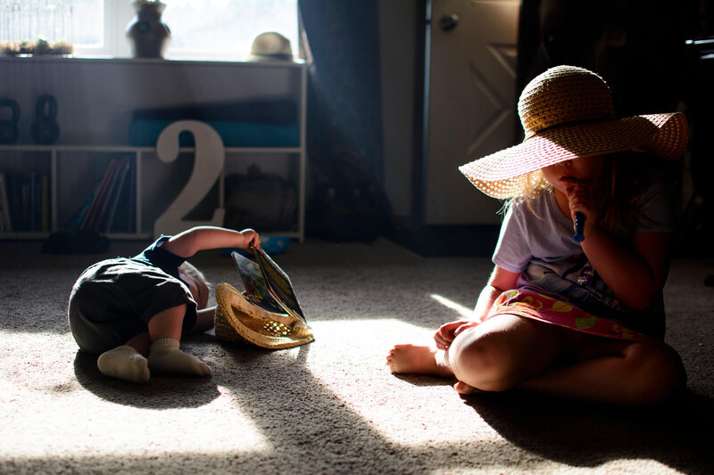 2 year old laying on floor, sister brushing teeth wearing hat - documentary family photography - wisconsin family photography