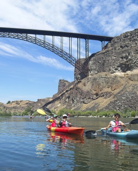 Two kayakers paddling along the Snake River beneath the iconic Perrine Bridge, surrounded by rugged canyon cliffs and clear blue skies